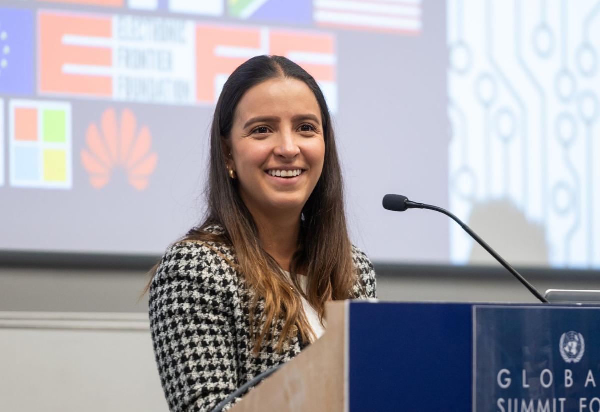 Female student is smiling at a lectern. Behind her some flags are displayed on a projector.